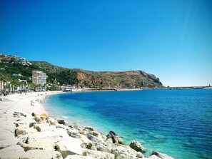Une plage côtière aux eaux bleues et limpides, avec un rivage rocheux et des bâtiments construits sur le flanc de la colline.
