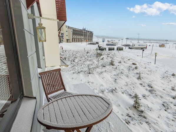 A balcony with a table and chair, overlooking a snowy beach area with parked cars and a building in the background.