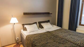 Quarto com cama, abajur, prateleira de madeira e janela com cortinas.