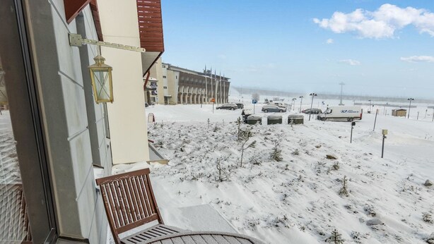 Ein Balkon mit Holzstuhl und Tisch, mit Blick auf eine verschneite Landschaft, in deren Hintergrund Parkautos und ein Gebäude zu sehen sind.
