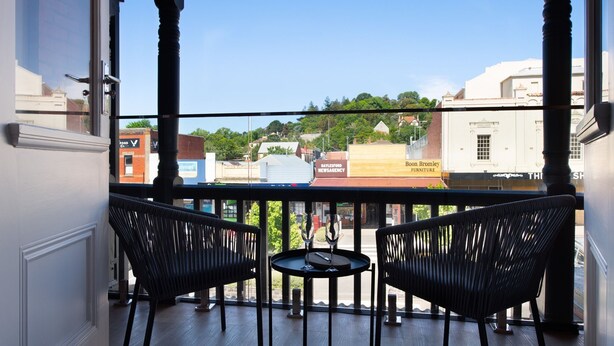A balcony with two black wicker chairs and a small round table, overlooking a street with various shops and buildings. - 1913 on Vincent Soaring ceilings and stunning Views (Daylesford)