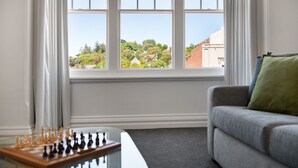 A room with a chessboard on a table, a grey sofa with a green pillow, and a window with a view of trees and buildings. - 1913 on Vincent Soaring ceilings and stunning Views (Daylesford)