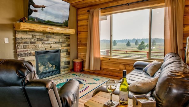A cozy cabin interior with a stone fireplace, leather sofas, a large window with a view, and a mounted television.