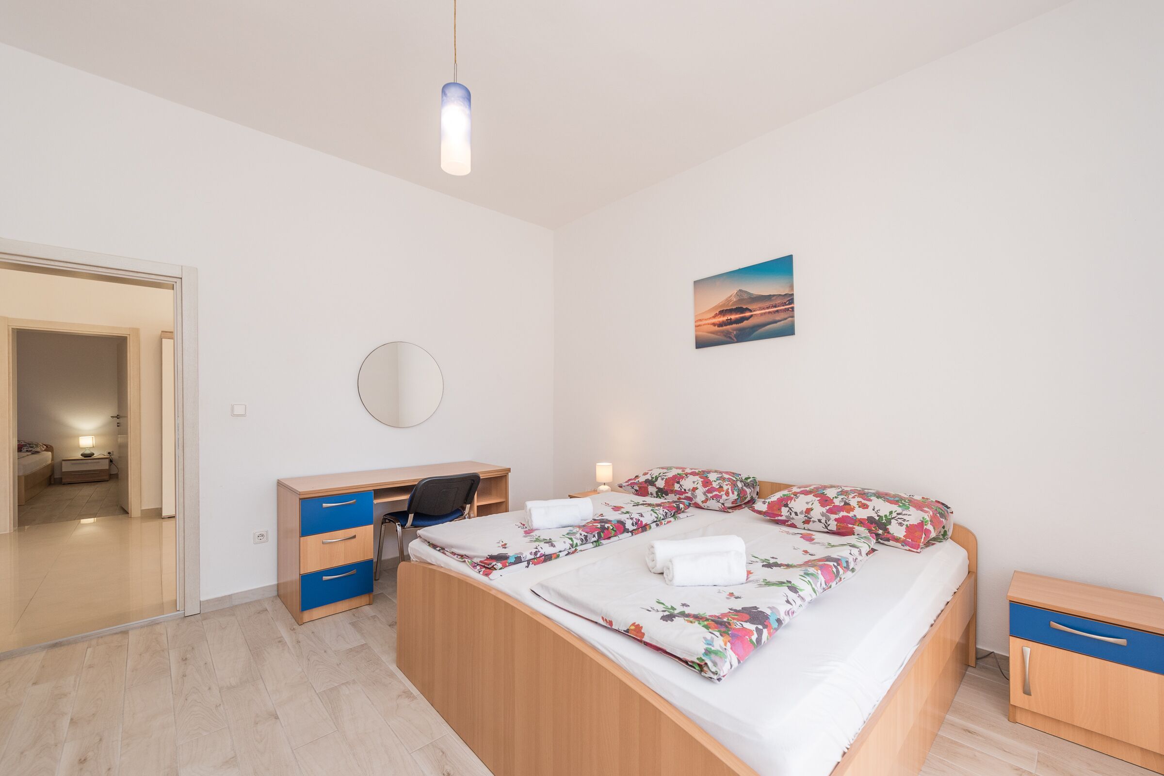 A bedroom with a bed, a desk, a chair, a bedside table, and a wall-mounted mirror.