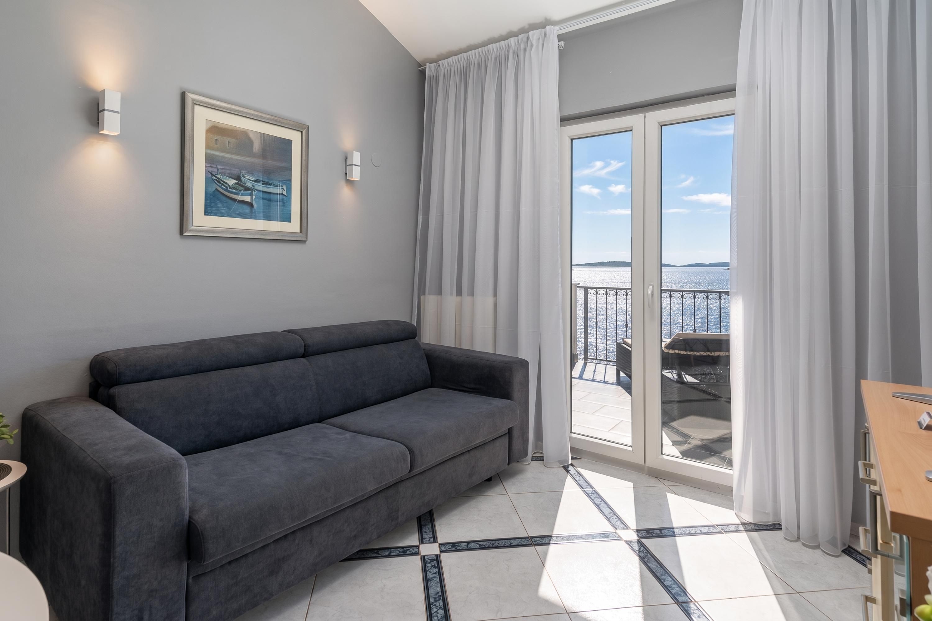 A modern living room with a grey sofa, a framed painting, and a balcony with a view of the sea.