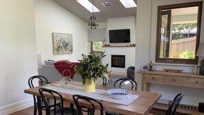 A dining area with a wooden table, black chairs, a vase with flowers, and a TV mounted on the wall. - Charming Luxury Self Contained Cottage. (Leura)