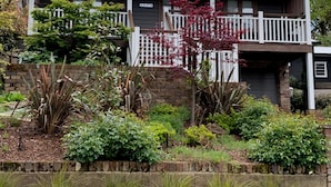 A two-story house with a covered porch, a brick wall, and a garden with various plants and shrubs. - Charming Luxury Self Contained Cottage. (Leura)