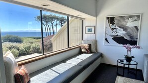 A room with a large window offering a view of the ocean and trees, a bench with cushions, a framed picture on the wall, and a small table with a vase of flowers. - Ocean & Golf Fairway view (Bodega Bay)