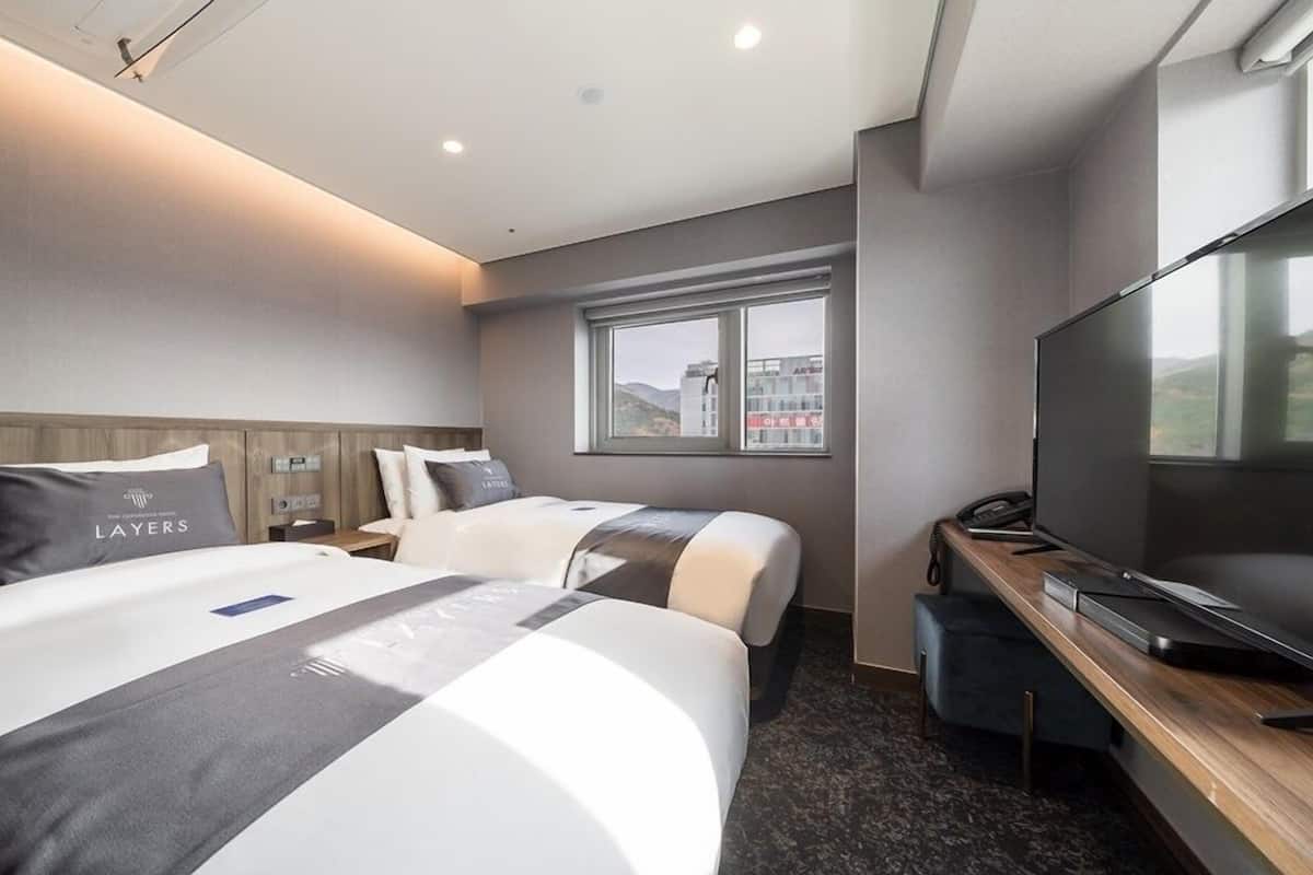 A hotel room with two beds, a desk, a television, and a window with a city view.