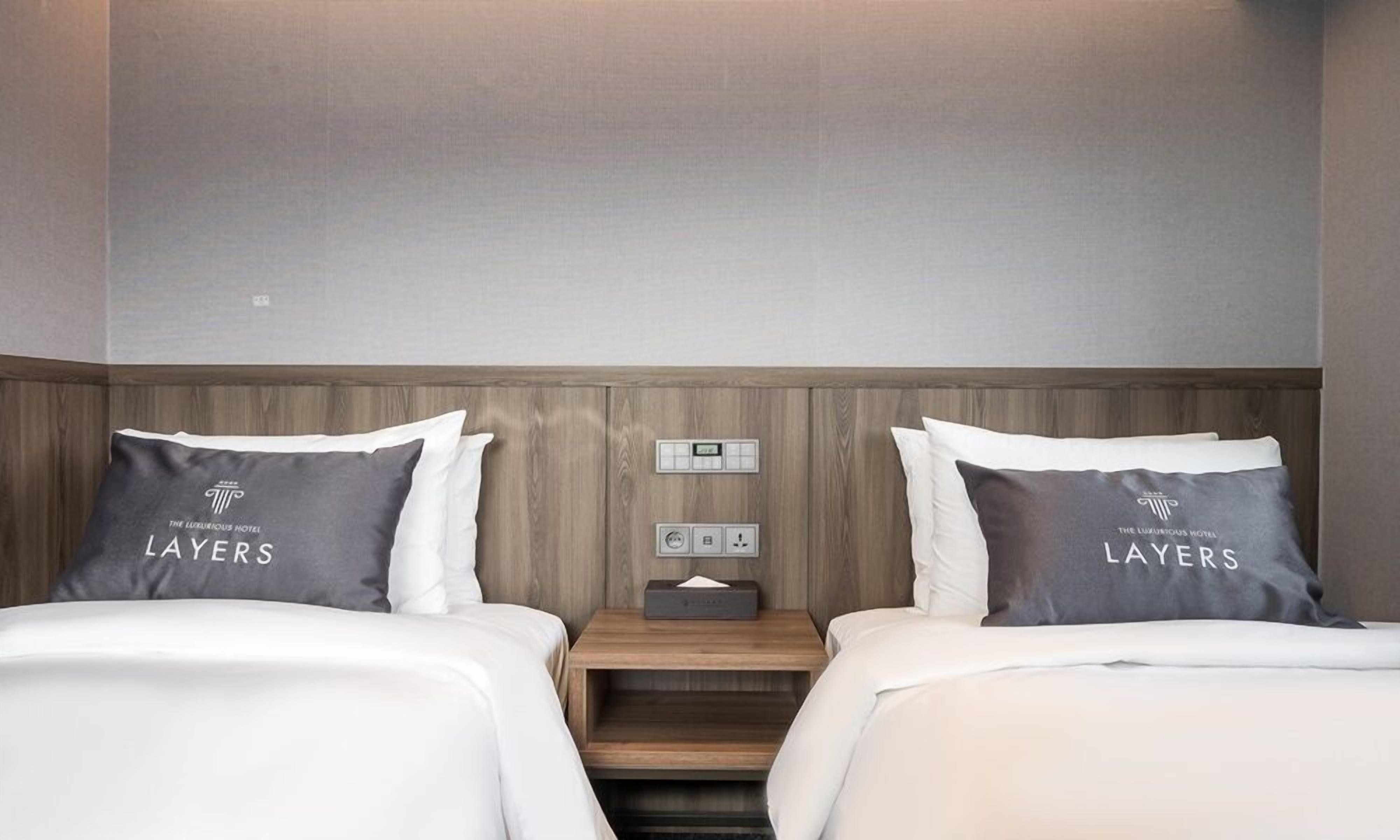 Habitación de hotel con dos camas, cada una con un almohadón que dice 'LAYERS'. Hay una mesita de noche con una lámpara y un panel de control.