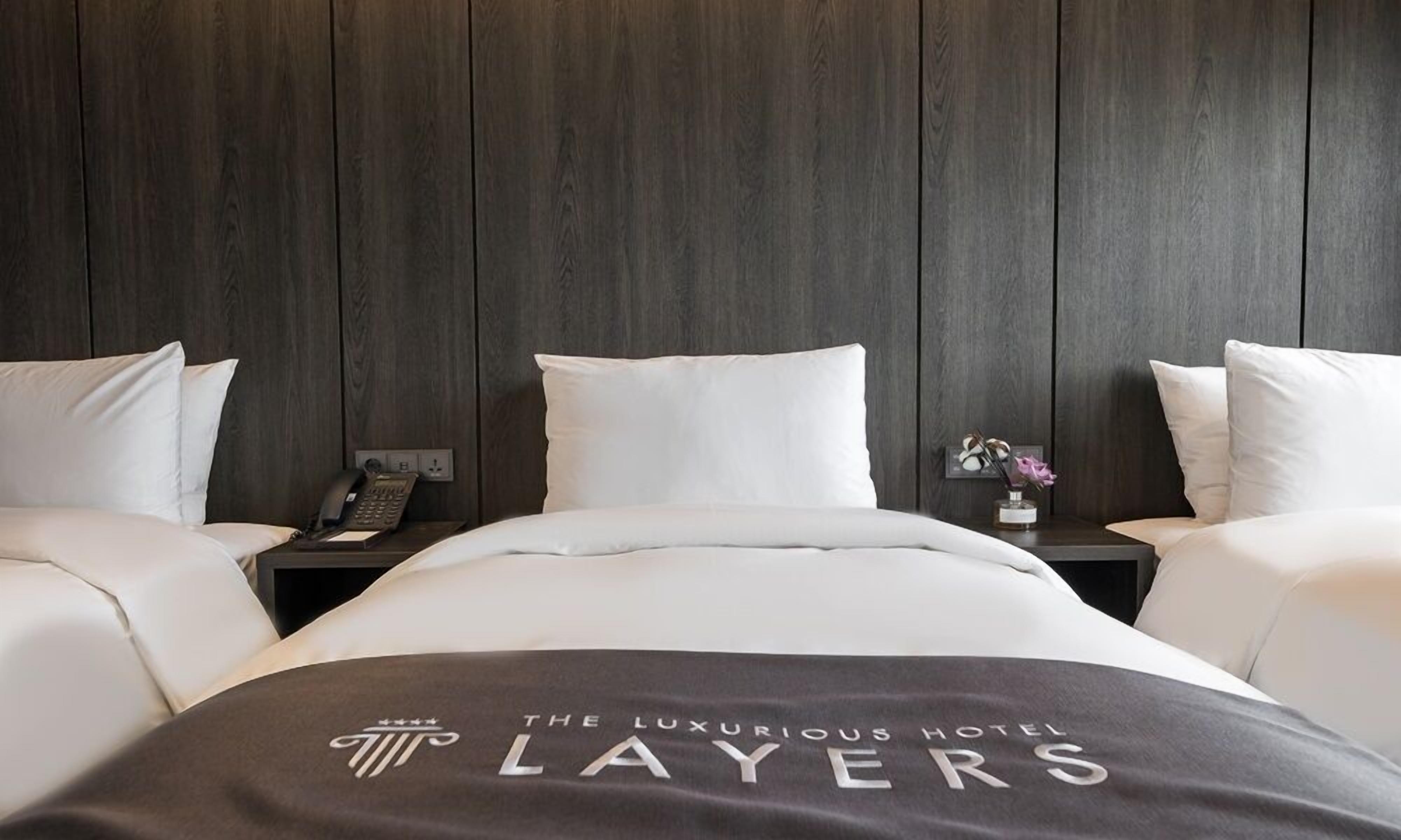 Habitación de hotel con pared de madera oscura, una cama con ropa blanca, dos mesitas de noche con un teléfono y un jarrón de flores, y una manta con el nombre del hotel.