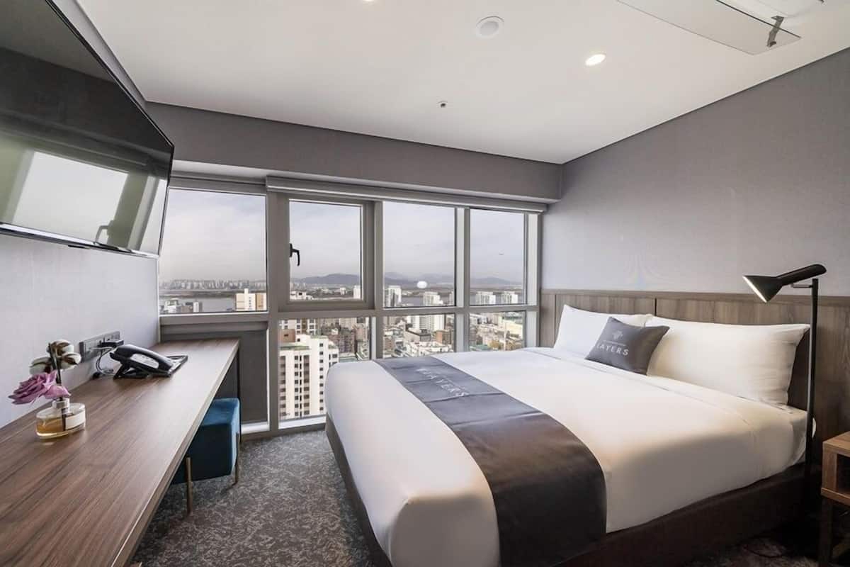 A hotel room with a large bed, a desk with a phone, a lamp, and a view of the city through large windows.