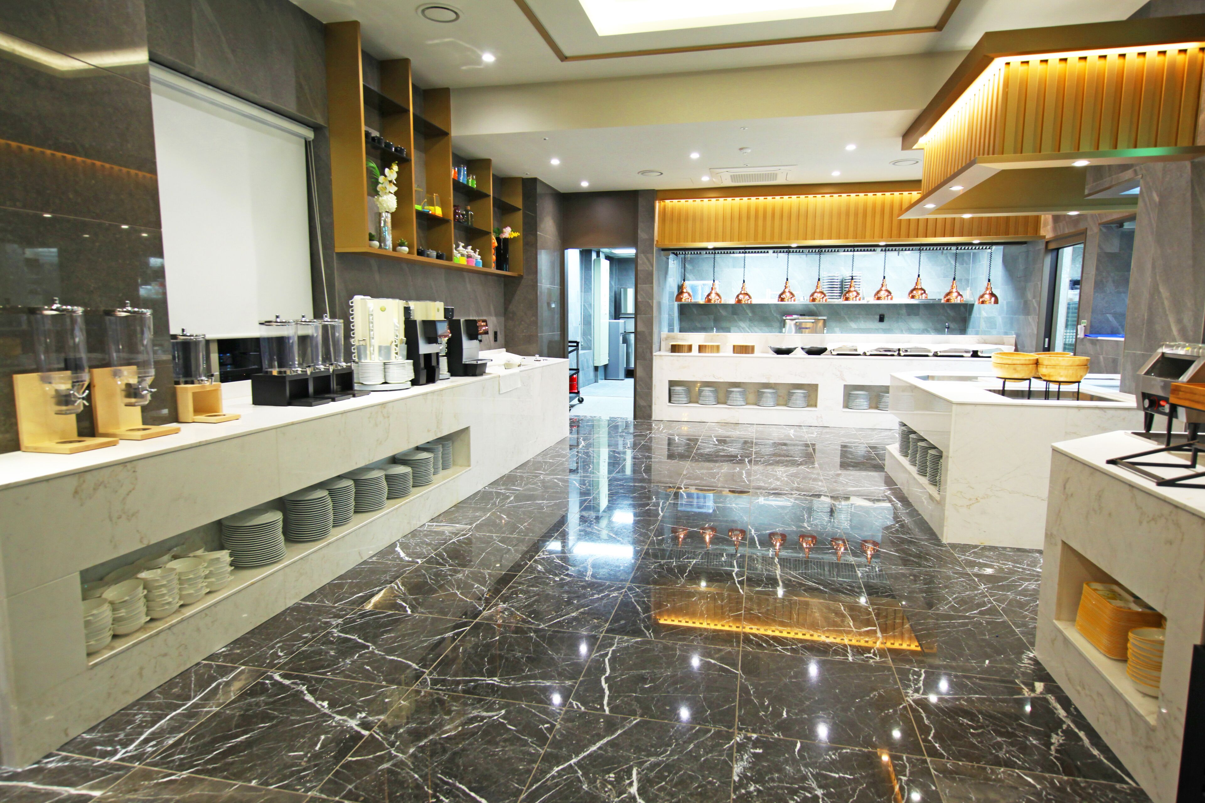 Um saguão de hotel moderno, com piso de mármore, um balcão de recepção e uma área de cozinha visível ao fundo.