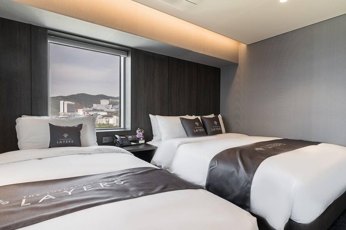 A hotel room with two beds, a large window with a city view, and a dark wood panel wall.