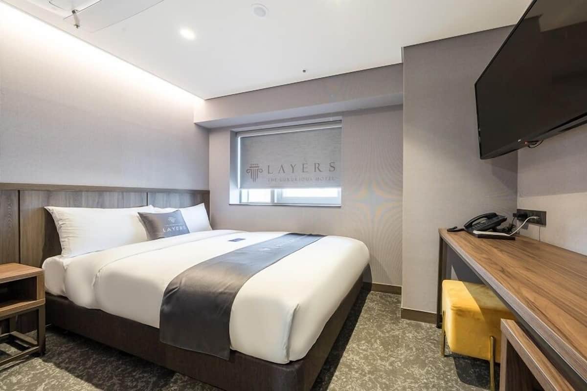 A modern hotel room with a large bed, a desk, a flat-screen TV, and a window with the hotel's name.