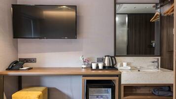 A hotel room with a flat-screen TV mounted on the wall, a wooden desk with a telephone, a coffee maker, and cups. There is a sink with a mirror above it, and a small stool in the corner.