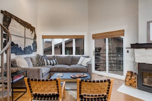 A modern living room with a sectional sofa, a coffee table, wicker chairs, a fireplace, and large windows with blinds.