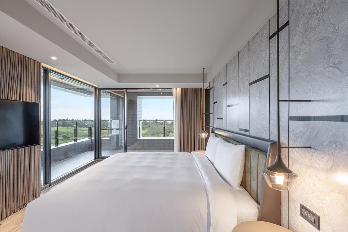 A modern hotel room with a large bed, a flat-screen TV, a balcony with a city view, and a marble wall.
