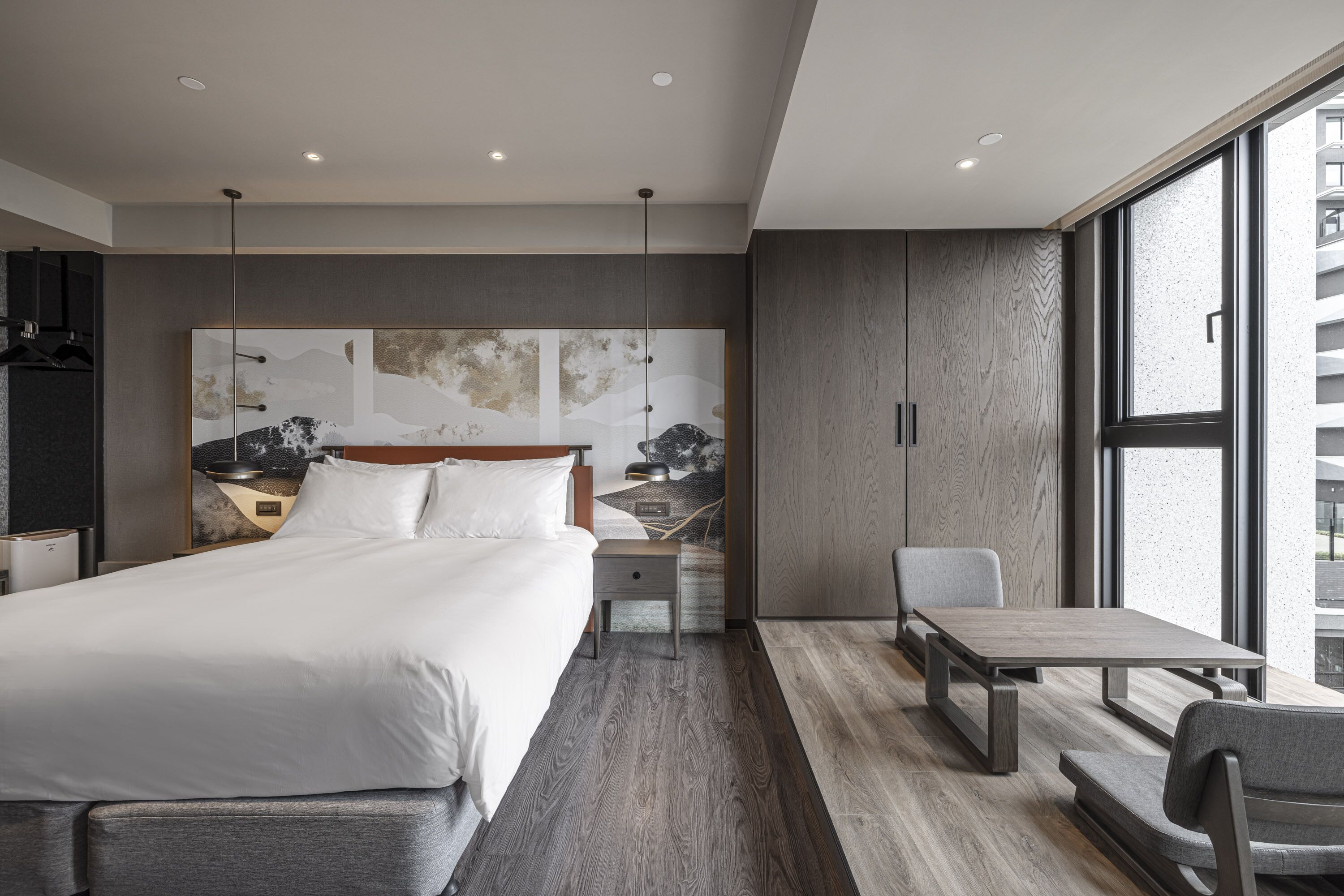 A modern hotel room with a large bed, a desk, and a chair. There is a large window providing a view of the city.