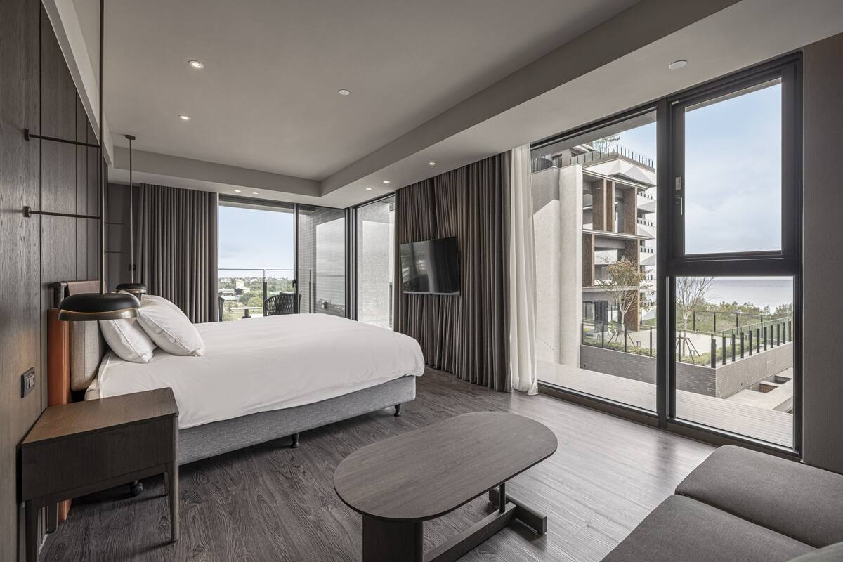 A modern hotel room with a large bed, a flat-screen TV, a small table, and a balcony with a view of the city.