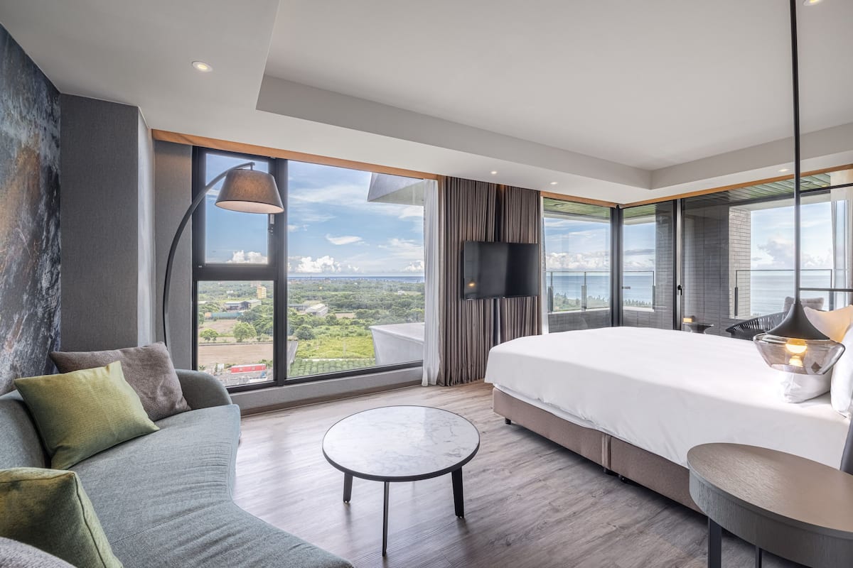 A modern hotel room with a large bed, a sofa, a round table, and a view of the city and greenery.