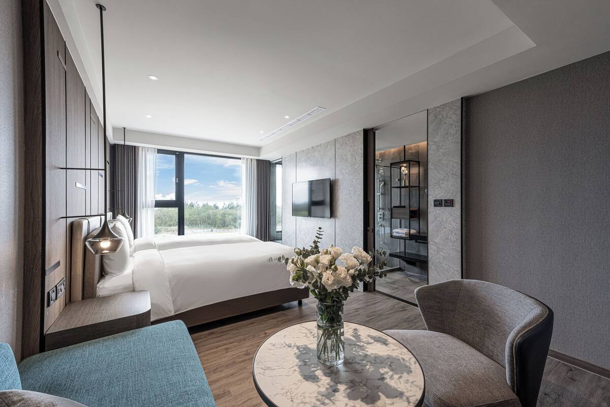 A modern hotel room with a large bed, a sitting area with a round table, and a view of the outdoors.