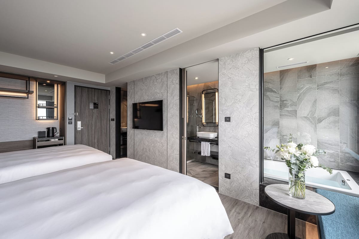 A modern hotel room with two beds, a TV, a bathroom with a large mirror, and a vase with flowers on a small table.