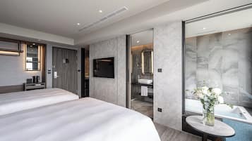 A modern hotel room with two beds, a TV, a bathroom with a large mirror, and a vase with flowers on a small table.