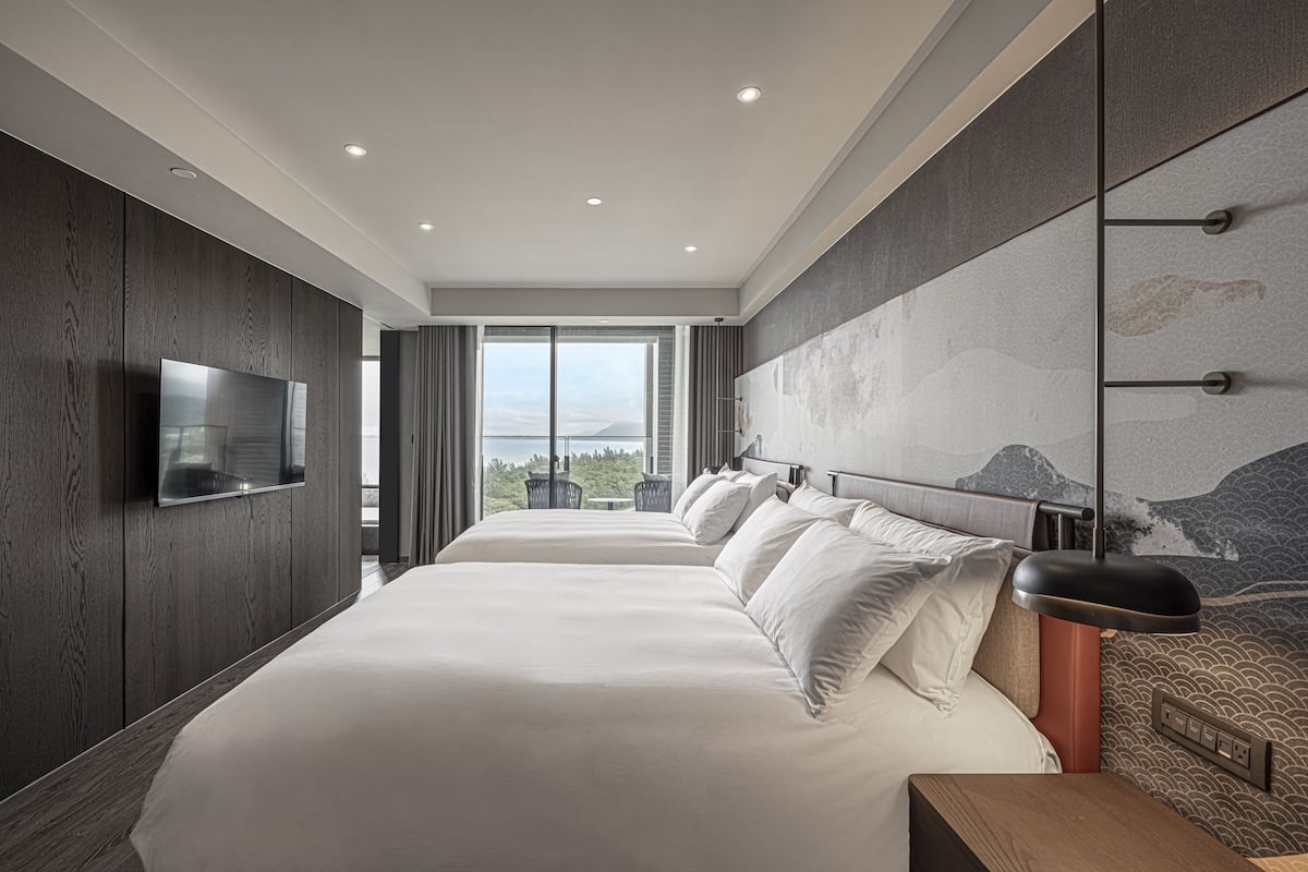 A modern hotel room with two large beds, a flat-screen TV, a large window with a view, and a wall-mounted light fixture.