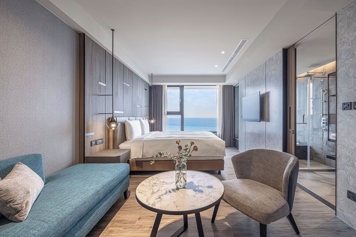 A modern hotel room with a large bed, a sofa, a round table, and a shower area.