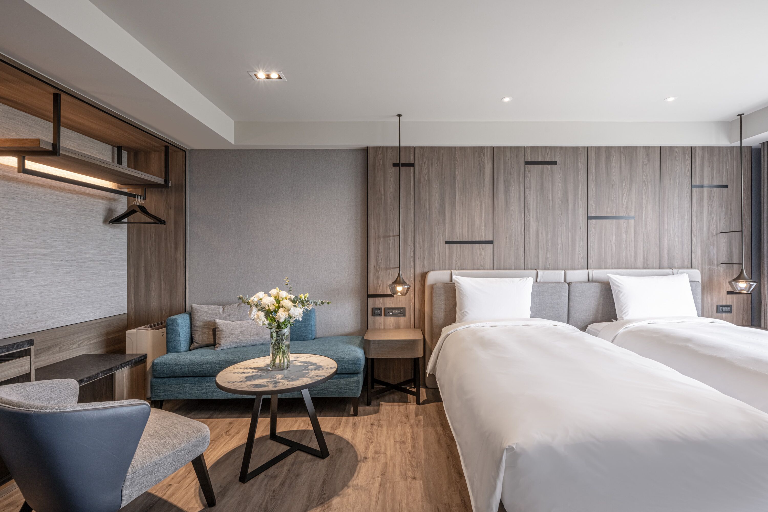 A modern hotel room with a large bed, a sofa, a small round table with a vase, and a desk with a chair.
