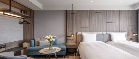 A modern hotel room with a large bed, a sofa, a small round table with a vase, and a desk with a chair.