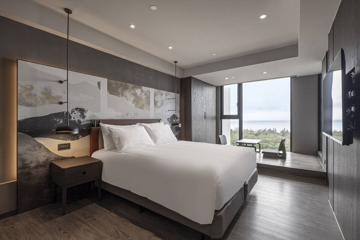 A modern bedroom with a large bed, a nightstand, a desk, and a view of the outdoors.