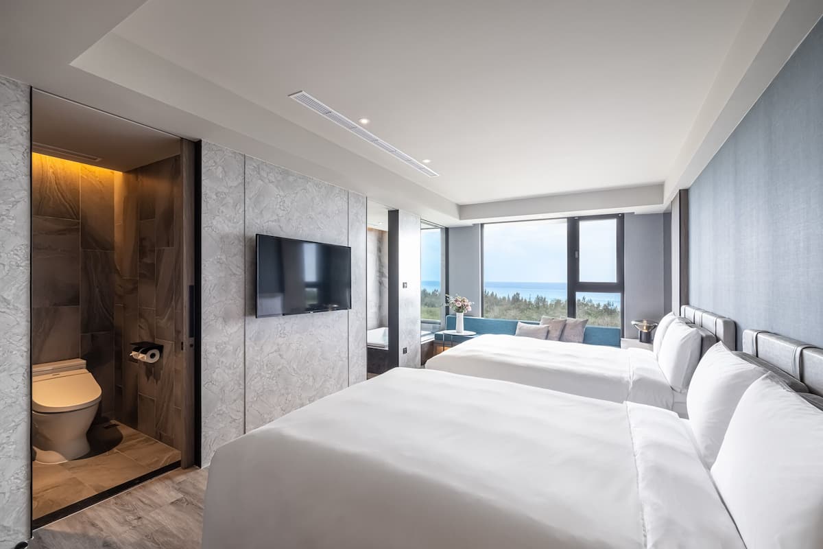 A modern hotel room with two beds, a TV, a bathroom with a toilet, and a view of the sea.
