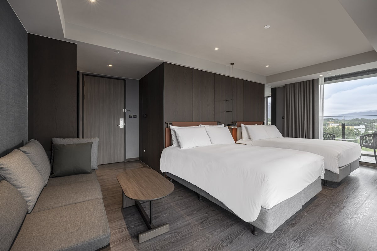 A modern hotel room with two beds, a sofa, a small table, and a large window offering a city view.