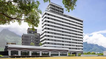 A modern multi-story building with a flat roof and large windows, surrounded by a landscaped area with trees and a view of mountains.