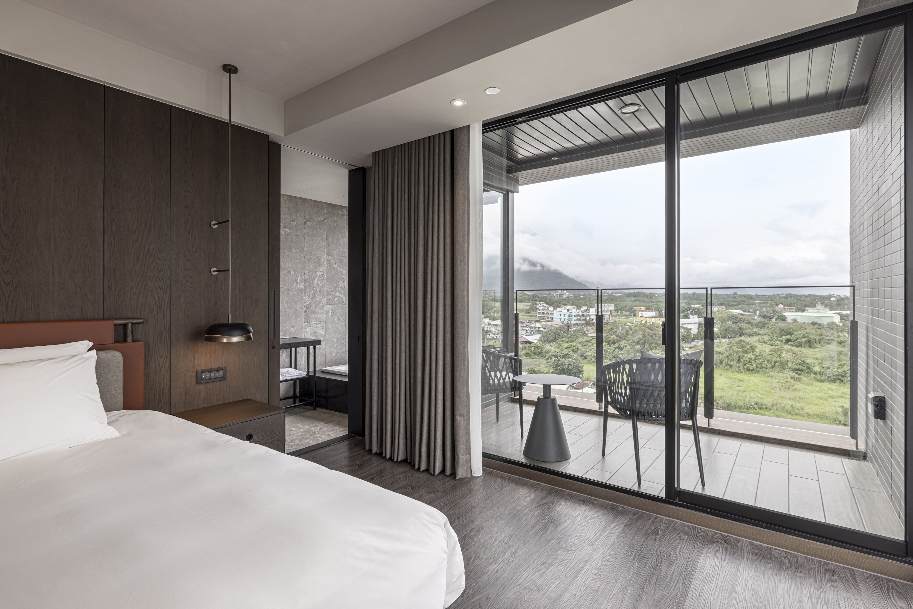 Quarto de hotel moderno com uma cama grande, uma escrivaninha pequena, uma varanda e vista para a cidade e montanhas.