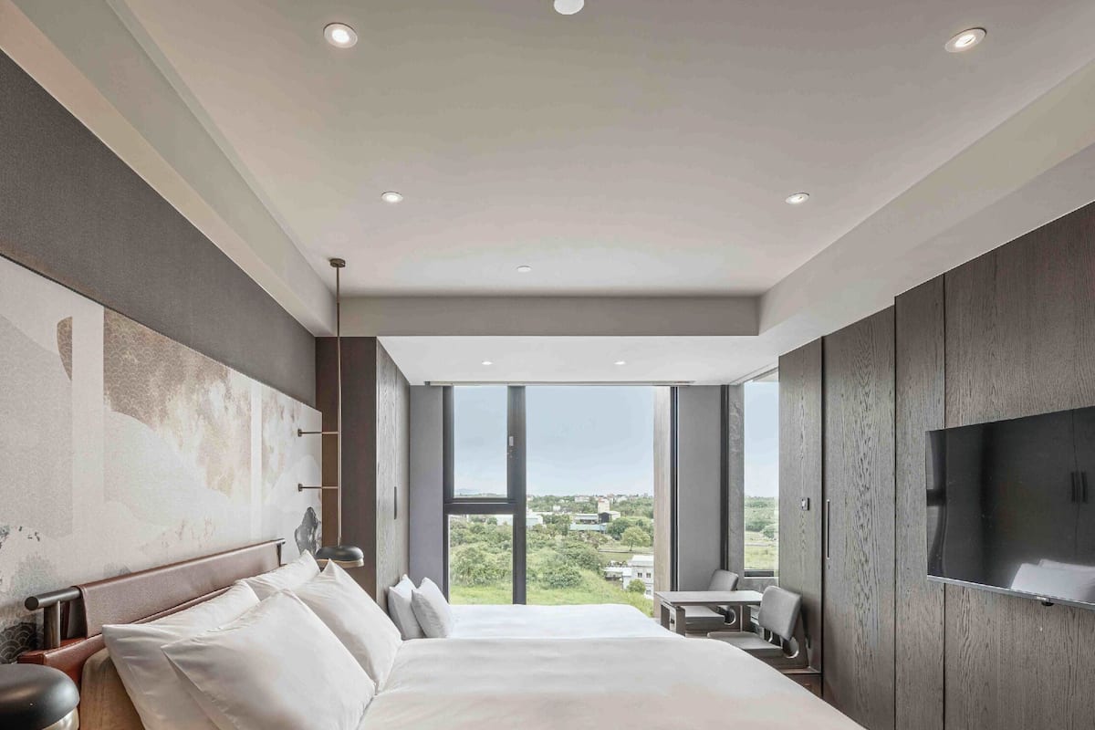 A modern hotel room with a large bed, a TV mounted on the wall, and a view of the city through large windows.