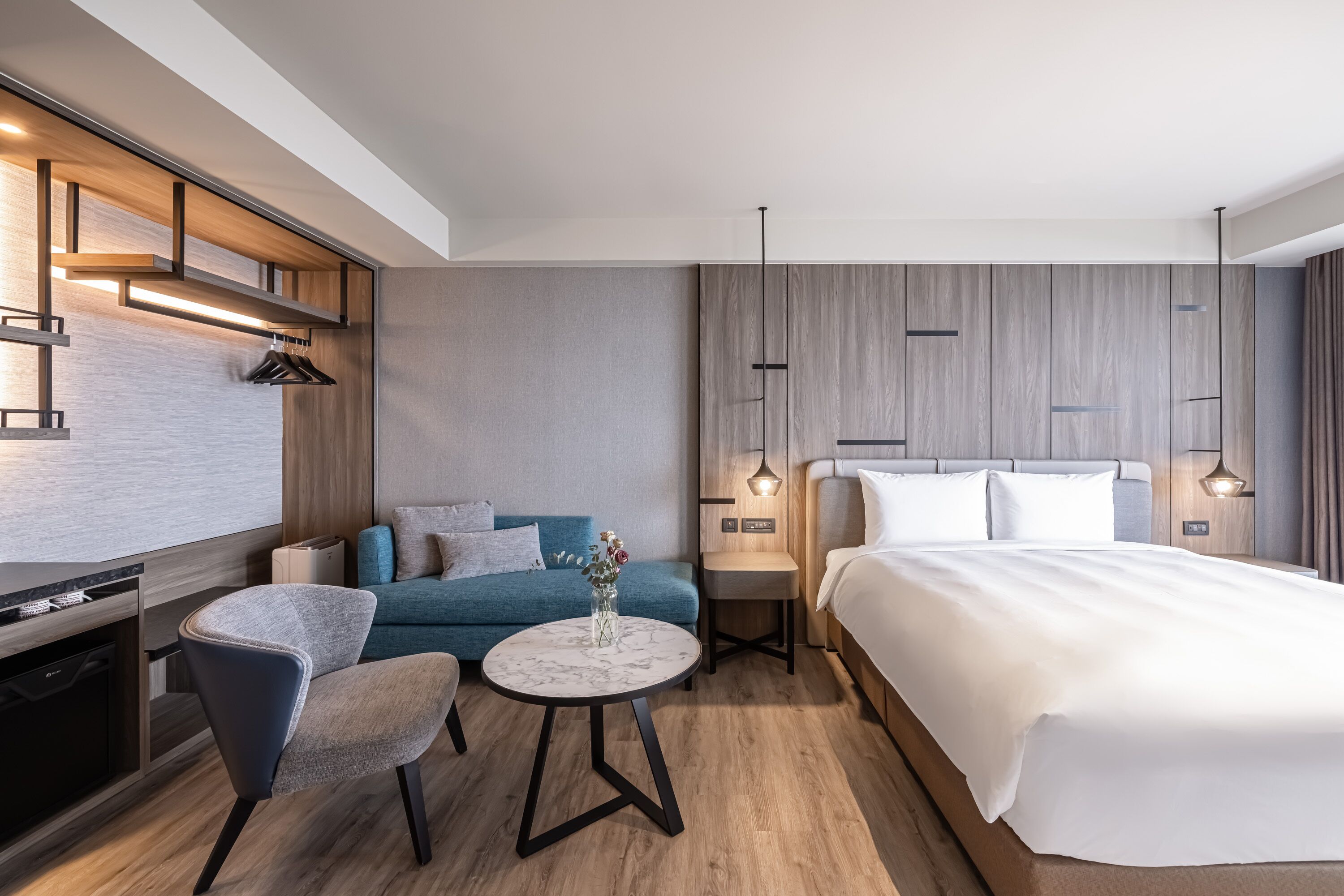 A modern hotel room with a bed, a sofa, a dining table, and a kitchen area.
