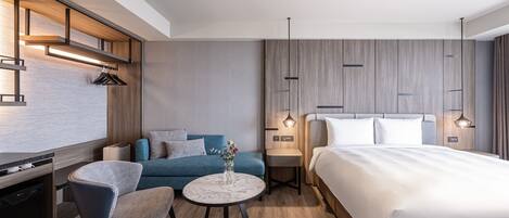 A modern hotel room with a bed, a sofa, a dining table, and a kitchen area.