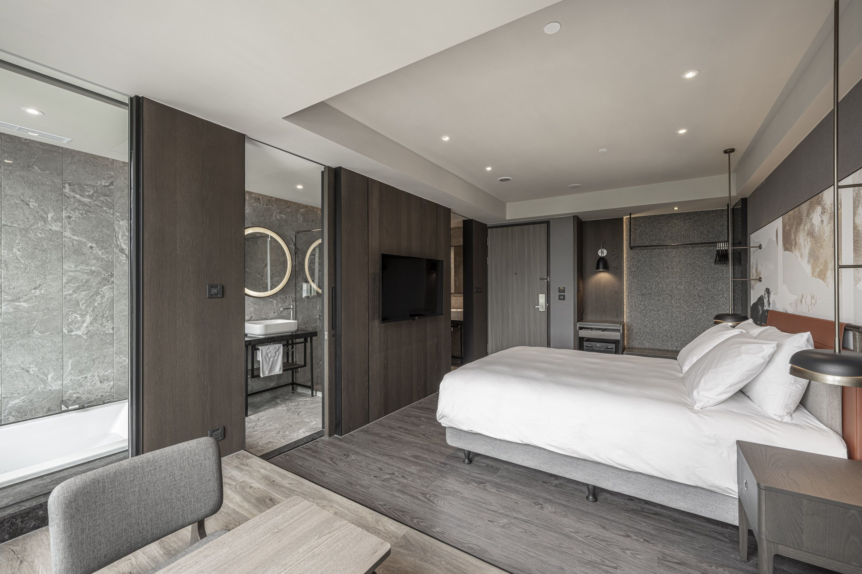 A modern hotel room with a large bed, a desk, a chair, a bathroom with a mirror, and a TV.