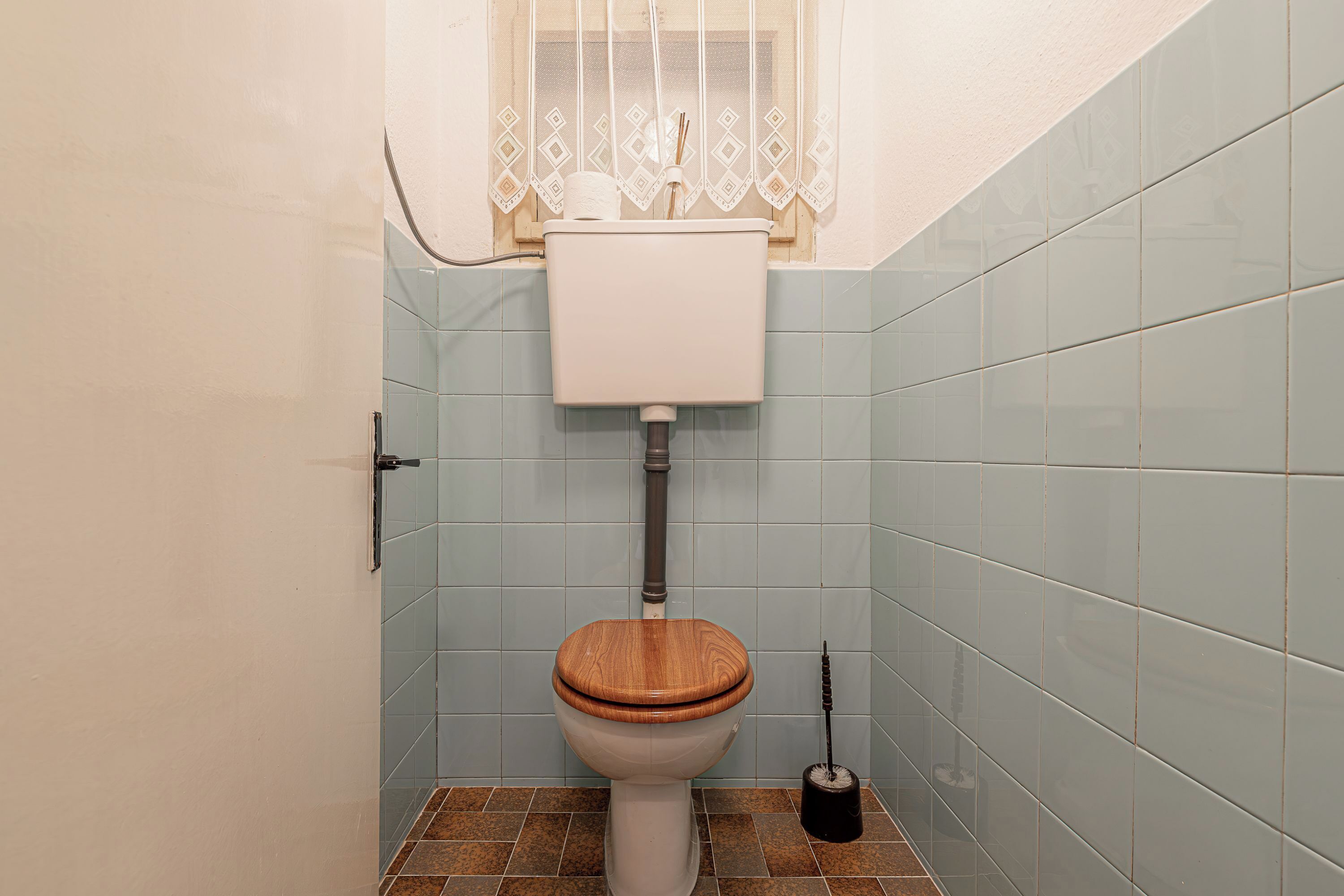 A bathroom with a toilet, a window with a decorative curtain, and tiled walls.