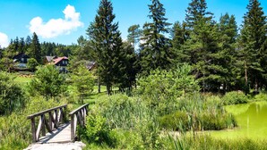 A wooden bridge leading to a pond surrounded by dense greenery and houses in the background. - Mržek House with Sauna (Bloke)