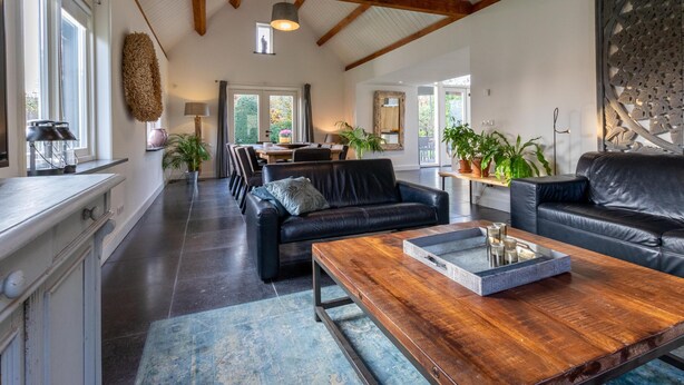 A spacious living area with a high ceiling, wooden beams, a dining table, black leather sofas, and potted plants. - Luxury holiday home with enclosed garden (Oosterhout)