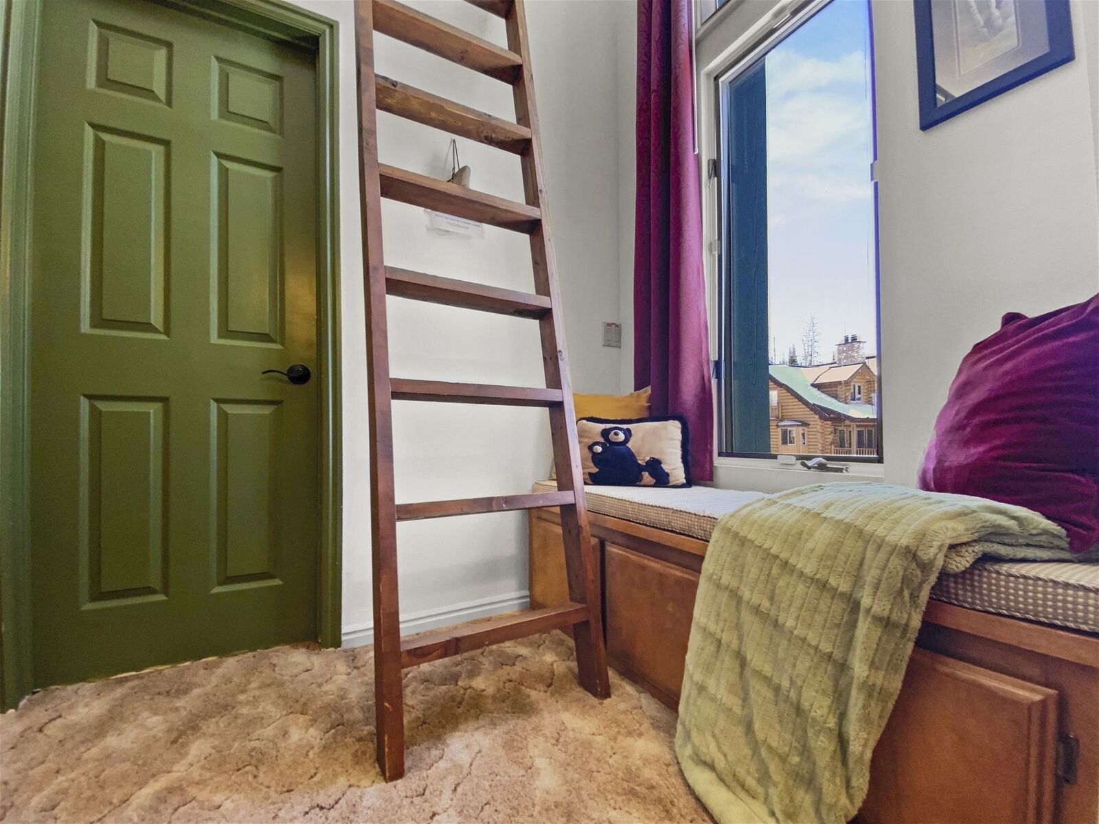 Una habitación con una escalera de madera apoyada en una pared, una ventana con vistas a edificios, una cama con un cojín morado y una puerta verde.