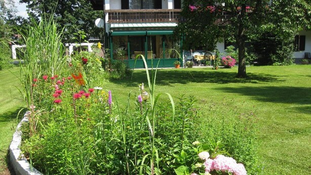 A garden with a variety of plants and flowers, a stone border, and a building with a green awning in the background. - Pension & Fewos Fuchs (Mauth)