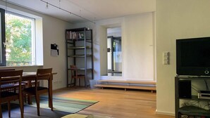 A modern living room with a wooden dining table, chairs, a bookshelf, a television, and a large window. - F80-Die FEINE Ferienwohnung (Regensburg)