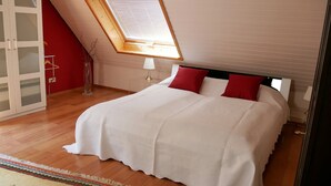 Un dormitorio con tragaluz, cama con ropa de cama blanca y almohadas rojas, suelo de madera, pared roja y un aparador de vidrio.