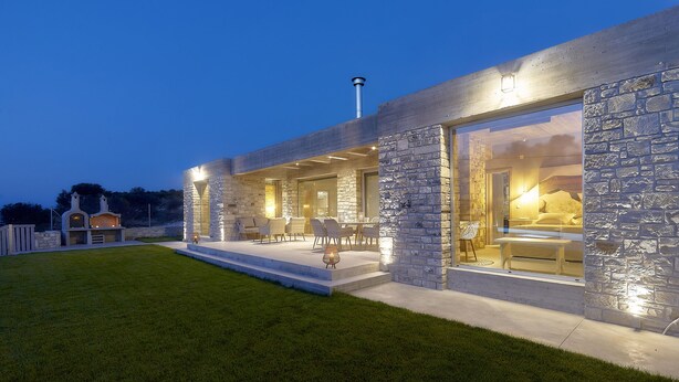 Una casa moderna con fachada de piedra, amplios ventanales y un área de estar exterior con sillas y una mesa.