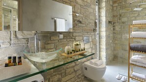 Un baño con pared de piedra, lavabo de vidrio, inodoro blanco y estante con artículos de aseo.